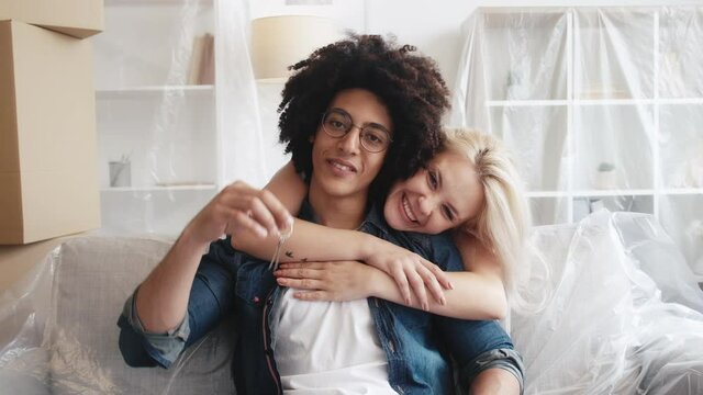 Couple relocation. Rent flat. Multiethnic family. Happy girlfriend embracing proud property owner guy showing keys of new place in living room interior with isolated furniture.