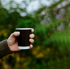 The man hand holding a blank paper cup in a wooded landscape background. Autumn lifestyle. Coffee black cup. Copyspace