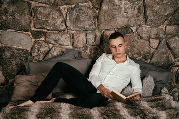 A young handsome man in a white shirt and pants lies on the bed reading a book before going to bed. An athletic guy in the bedroom.