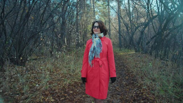 A girl in a red raincoat walks through the autumn forest. Brunette in black glasses on a forest path. She breathes in the autumn air and enjoys the new day. Beautiful country autumn landscape.