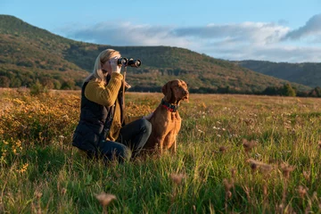Foto op Canvas Young female hunter using binoculars for bird spotting with hungarian vizsla dog by her side, out in a meadow on a beautiful sunny autumn evening. Hunting with a hunting dog. © andreaobzerova