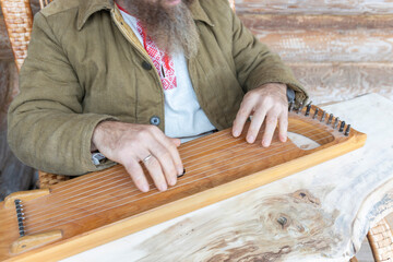 A man in ethnic clothes plays an ancient musical instrument. A bearded man, a forester has built a house and is playing a psaltery.
