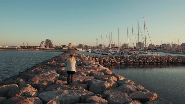 A girl standing on the rocks looking at the yachts and the city in the middle of the beach at sunrise, La Grande Motte , Montpellier - France