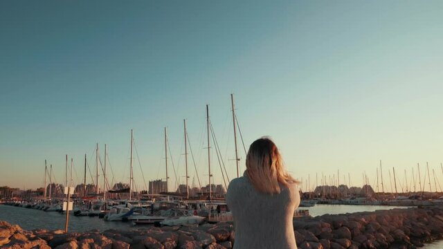 A girl is standing on the rocks looking at the yachts, the city in the middle of the beach, using her phone to take pictures at sunrise