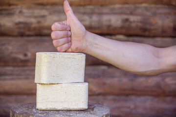 Fuel briquettes from pressed sawdust against the background of a log wall. Hand with a raised thumb.