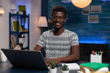 Portrait of african american man looking into camera while sitting at desk in living room late at night. Freelancer browsing management information on laptop computer working remote from home © DC Studio