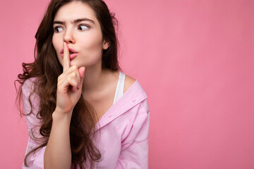Closeup of young emotional attractive wavy-haired brunette woman with sincere emotions wearing casual pink shirt isolated over pink background with copy space and showing silence gesture with finger