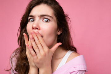 Closeup of young shocked astonished beautiful curly brunet female person with sincere emotions wearing casual pink shirt isolated over pink background with free space and covering mouth with hands