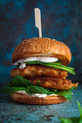 Fish burger on gray plate, dark blue background. Cod fish in batter with mayonnaise and spinach...