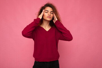 Portrait of young emotional beautiful brunette curly woman with sincere emotions wearing trendy pink pullover isolated on pink background with copy space and having headache