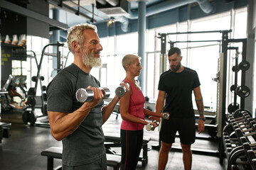 Approachable trainer supporting elderly couple while gym workout