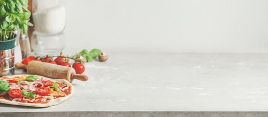 Hausgemachter Pizzahintergrund mit hölzernem Nudelholz, Mehl und Basilikumblättern auf grauem Küchentisch auf weißem Wandhintergrund. Italienisches Essen zu Hause kochen. Vorderansicht mit Kopierbereich.