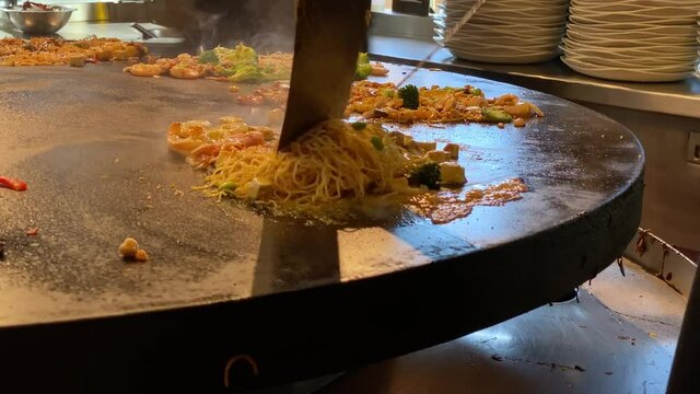 Close Up of Chef Stir Frying Noodles, Vegetables and Seafood with Spatulas on a Large Stove at a Mongolian Grill