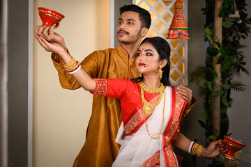 Portrait of Indian man dressed in kurta pajama with beautiful Indian woman wearing traditional Indian saree, gold jewellery and bangles, holding dhunachi in hands.