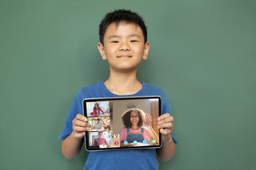 Smiling asian boy holding tablet for video call, with diverse elementary school pupils on screen