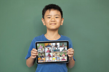 Smiling asian boy holding tablet for video call, with diverse elementary school pupils on screen