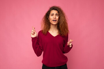 Photo shot of young attractive brunette curly woman with sincere emotions wearing trendy pink sweater isolated over pink background with empty space and holding fingers crossed for good luck. Gesture