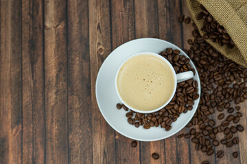 A cup of creamy coffee with roasted coffee beans next to it, wooden background