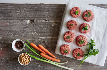 Freshly prepared homemade raw vegan burger patties placed on white parchment paper with some ingredients: spring onions, carrots, chickpeas, parsley and pepper on a wooden table. Meat alternative.