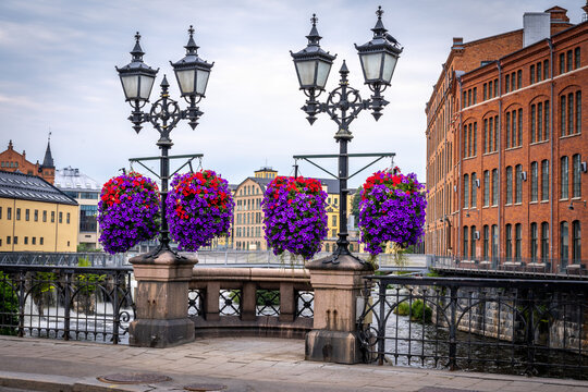 Beautiful cityscape view of purple flowers in flowerpot hanging from vintage lampposts. Industrial buildings in the background in Norrkoping Sweden.