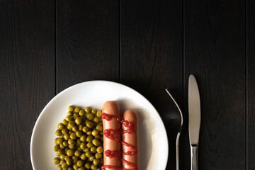 Sausages with ketchup, a side dish of peas on a dark background. Top view. White plate and stainless steel fork. Copy space.