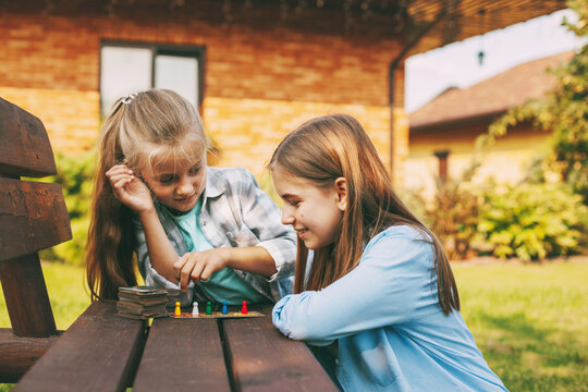 Two sisters laugh and play a wooden board game with colorful blue, red, green and yellow chips outdoors outside their home.