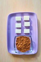 A glass cup with cocoa powder and six slices of white sugar lie on a purple plate. Top view