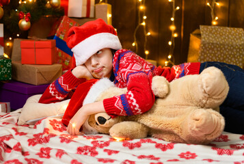 Child girl posing and dreaming in new year or christmas decoration. Holiday lights and gifts, Christmas tree decorated with toys. She's wearing a red sweater and a Santa helper hat