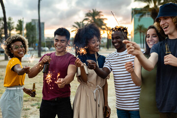 Multicultural united group of young friends having party together with sparkles while drinking beer outdoor - Community, friendship, unity and people celebrating concept - Focus on man on the corner