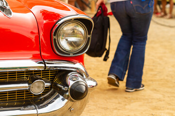 Front detail of fender and headlight of red chevrolet with people out of focus in the background in...