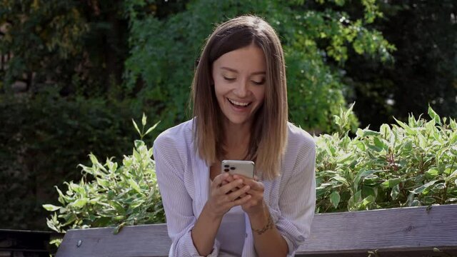 Happy young woman sitting on the bench and looking at the phone. Happy reaction and good news emotions. 