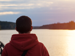 A man in an orange hoodie looks at a cloudy autumn sunset by the lake. Lifestyle, rest and freedom