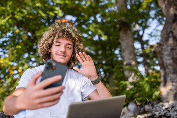 An Afro man sits in nature and uses a smartphone and a laptop for an online meeting