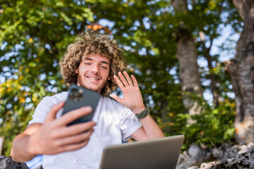 An Afro man sits in nature and uses a smartphone and a laptop for an online meeting