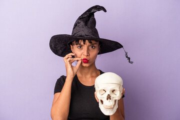 Young mixed race woman disguised as a witch holding a skull isolated on purple background  with fingers on lips keeping a secret.