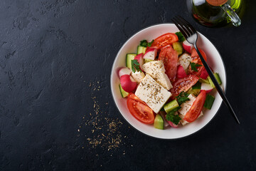 Radish, cucumber, tomato, pepper and feta cheese with spices pepper and olive oil in white bowl on black slate, stone or concrete background. Healthy food concept. Top view.