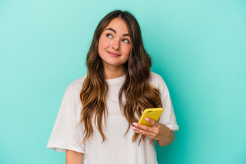 Young caucasian woman holding a mobile phone isolated on blue background dreaming of achieving...