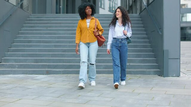 Two cheerful diverse women walking down stairs and laughing while talking. Female best friends strolling in town and having nice time together