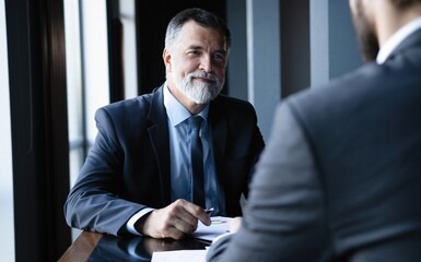 Portrait of a confident mature businessman sitting at table in office, meeting with colleague.