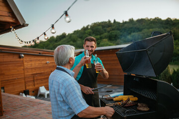 Young man and his gray hair father toasting with bottle of beer while roasting meat on barbecue...