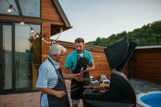 Senior man holding bottle of beer, and talking with his son while barbecuing in the backyard