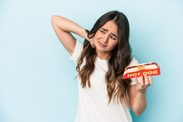 Young caucasian woman holding a hot dog isolated on blue background touching back of head, thinking and making a choice.