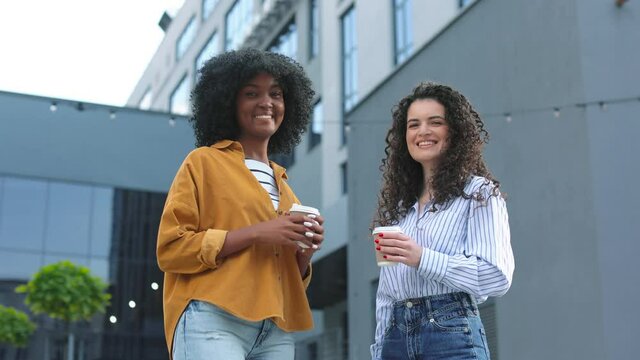 Portrait of multi-ethnic young women standing in city with coffee-to-go in hands. Beautiful joyful caucasian and multiracial women speaking outdoors on street. Conversation concept
