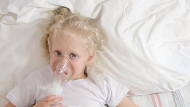 cute little girl with curly blond hair lies in bed, sick five-tape girl in bed makes inhalation with a nebulizer, treatment of bronchitis and lung disease in children