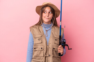 Young caucasian fisherwoman holding a rod isolated on pink background confused, feels doubtful and unsure.