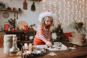 little girl in a Santa hat and red striped pajamas cooks a Christmas cake in a beautiful kitchen decorated with a garland. merry Christmas and happy New Year and Holidays. hygge. High quality photo