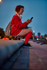 Smiling female skateboarder relaxing at the quay and using cellphone