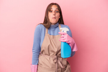 Young caucasian cleaner woman holding spray isolated on pink background shrugs shoulders and open eyes confused.