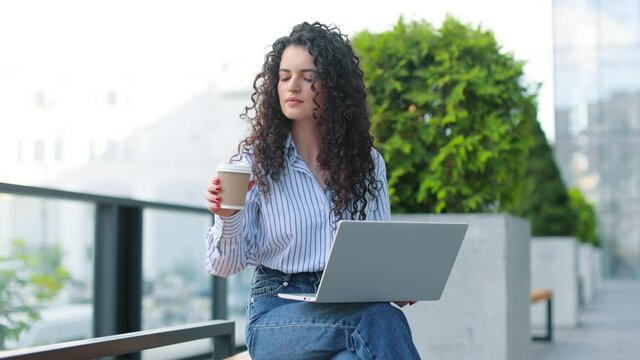 Good looking caucasian woman having free time while sitting on the bench with a laptop at her knees. Outdoors