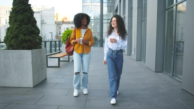 Two pretty female friends in nice outfits walking through the city street and talking joyfully. Beautiful friends women stepping outdoor and sharing secrets or gossips